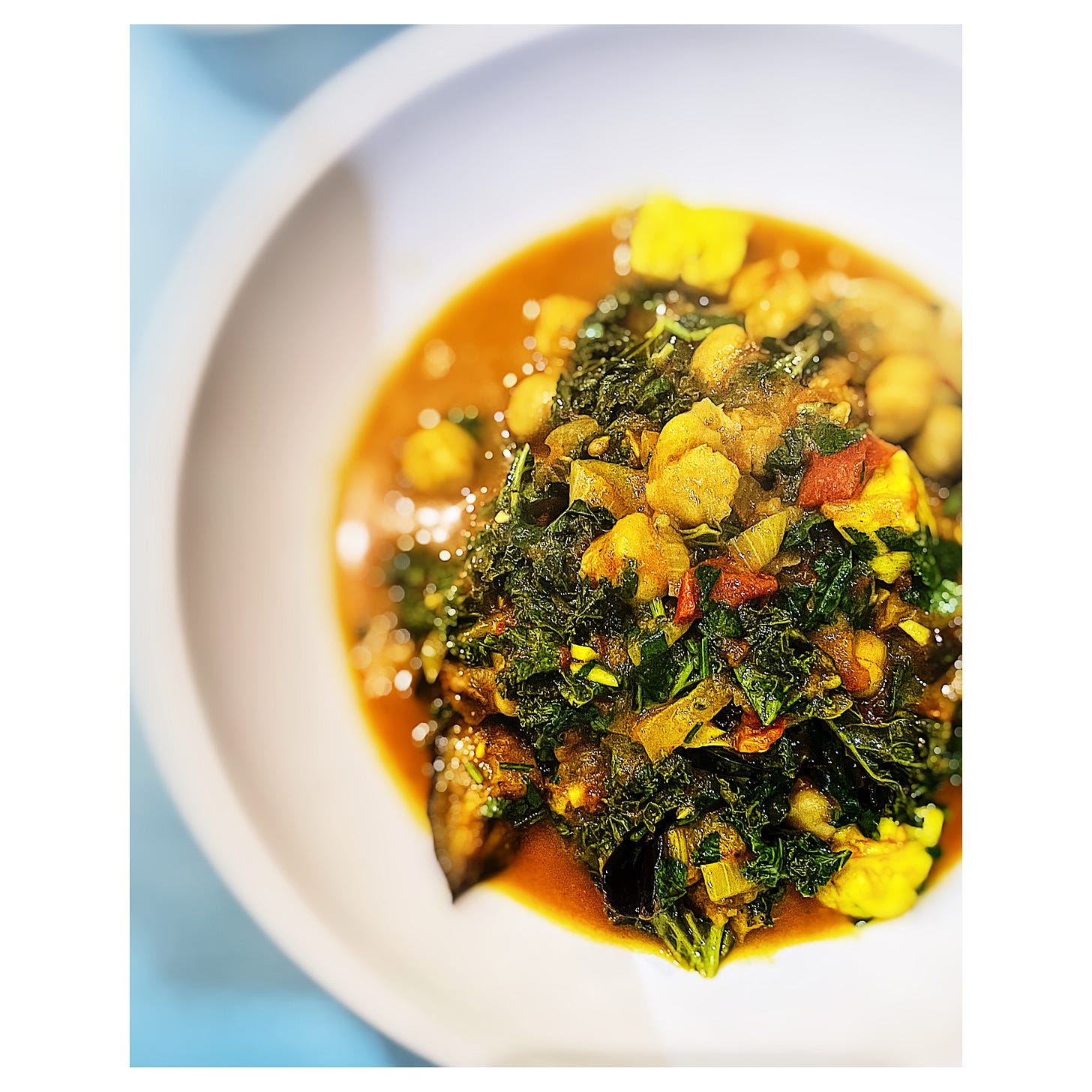 An over head photo of part of a bowl holding a serving of curry - there is a dark orange sauce with chickpeas, pieces of aubergine, cubes of paneer and chopped dark greens in