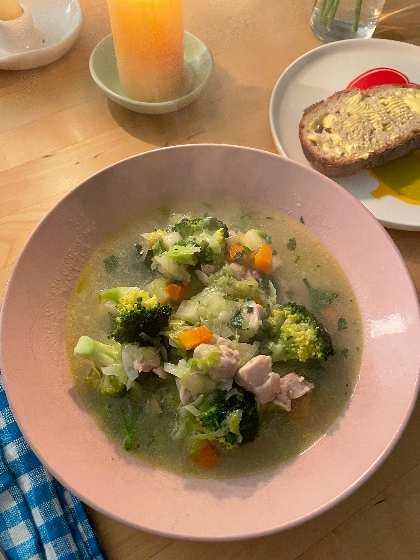 Bowl of chicken and vegetable soup in a pink bowl. It's steaming and there's a piece of buttered bread nearby and candles to light the table.
