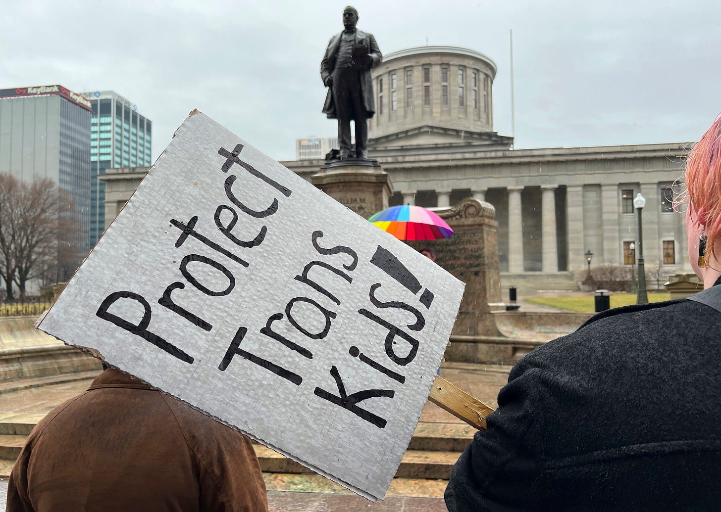 Protester holding a sign that says ''Protect Trans Kids!'' stands outside of the Ohio Statehouse .
