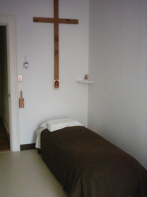A small bedroom, called a cell, where the sisters used to sleep in Brooklyn. It’s sparse, containing a twin bed covered by a dark brown blanket. A large wooden cross is on the wall over the bed. 