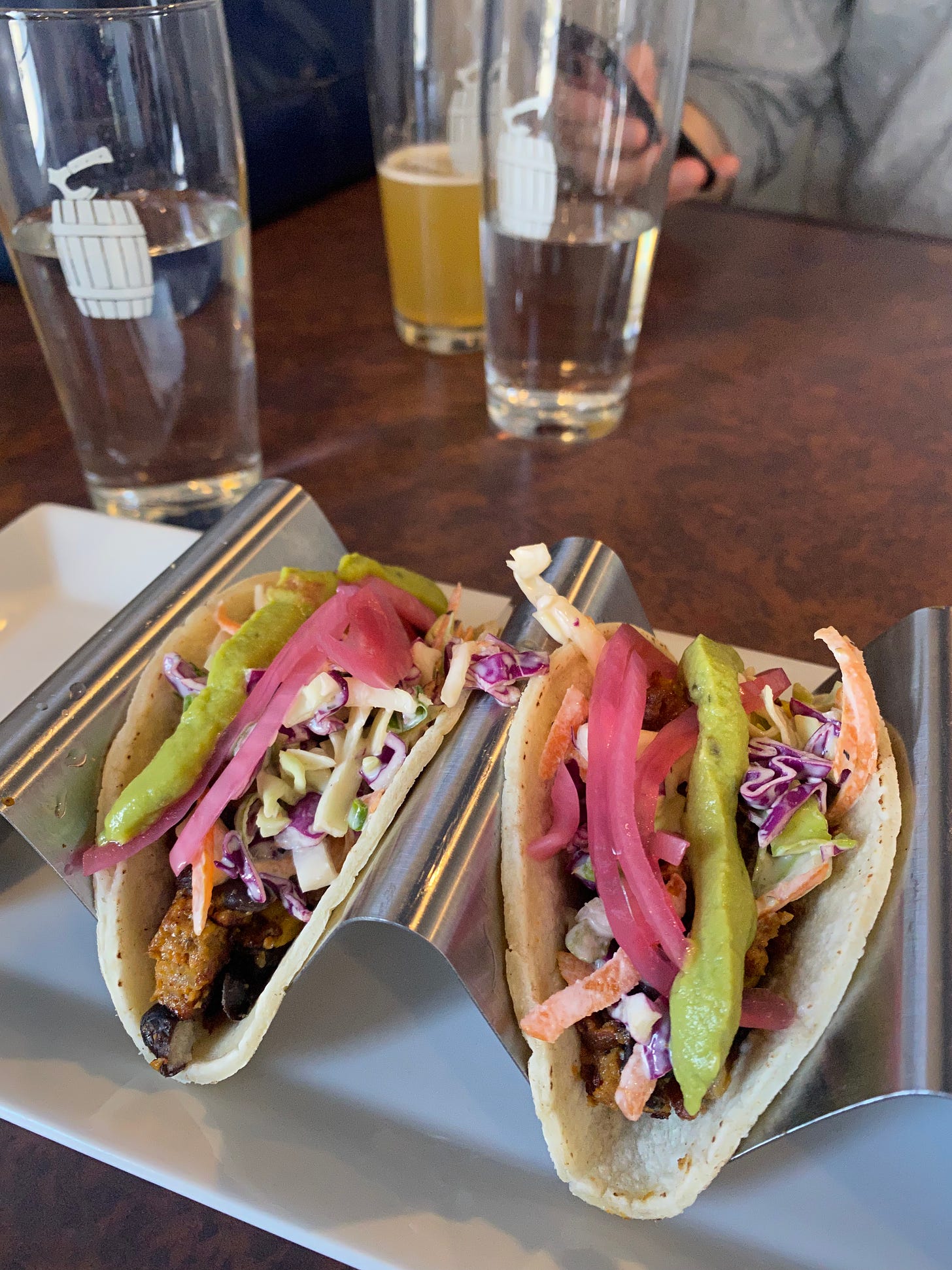 Two tacos in a metal taco stand on a white plate. The tacos have beans and fried veggies on the bottom and are topped with slaw, pickled red onion, and avocado cream.