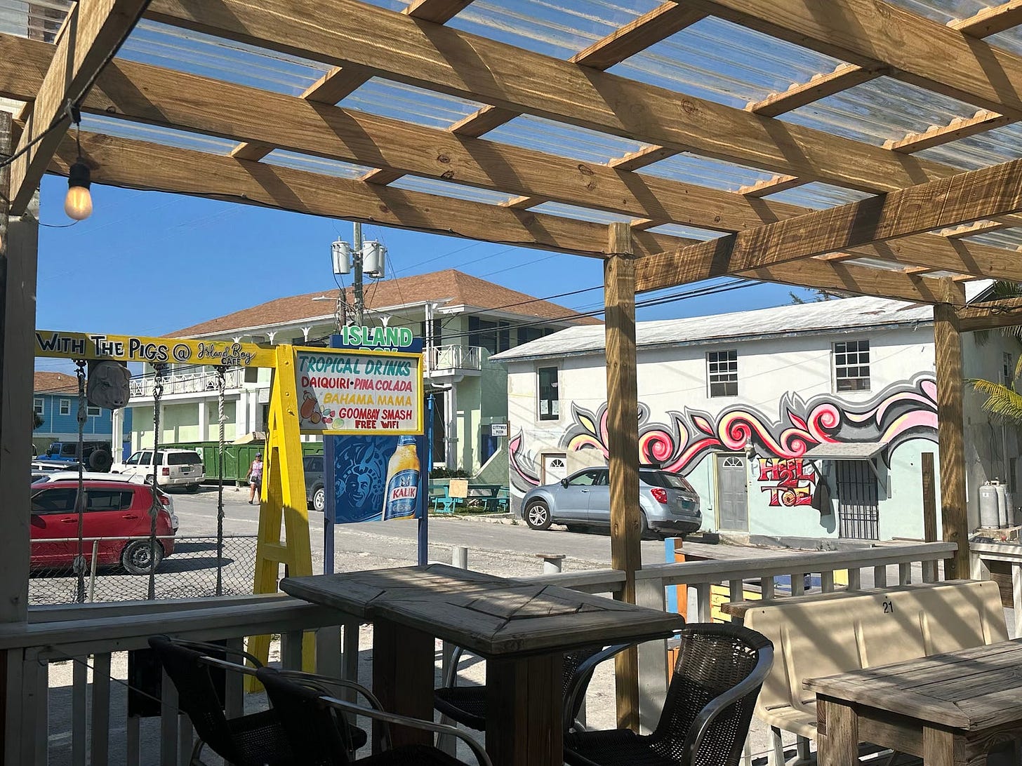 Wood pergola over picnic tables and chairs. A yellow sign — “Island Bar” and has list of a few cocktails. A colonial-style building is at the back of the parking lot with another plain white two-story building. There is pink and grey street art on the white building, that says “Heel 2 Toe”