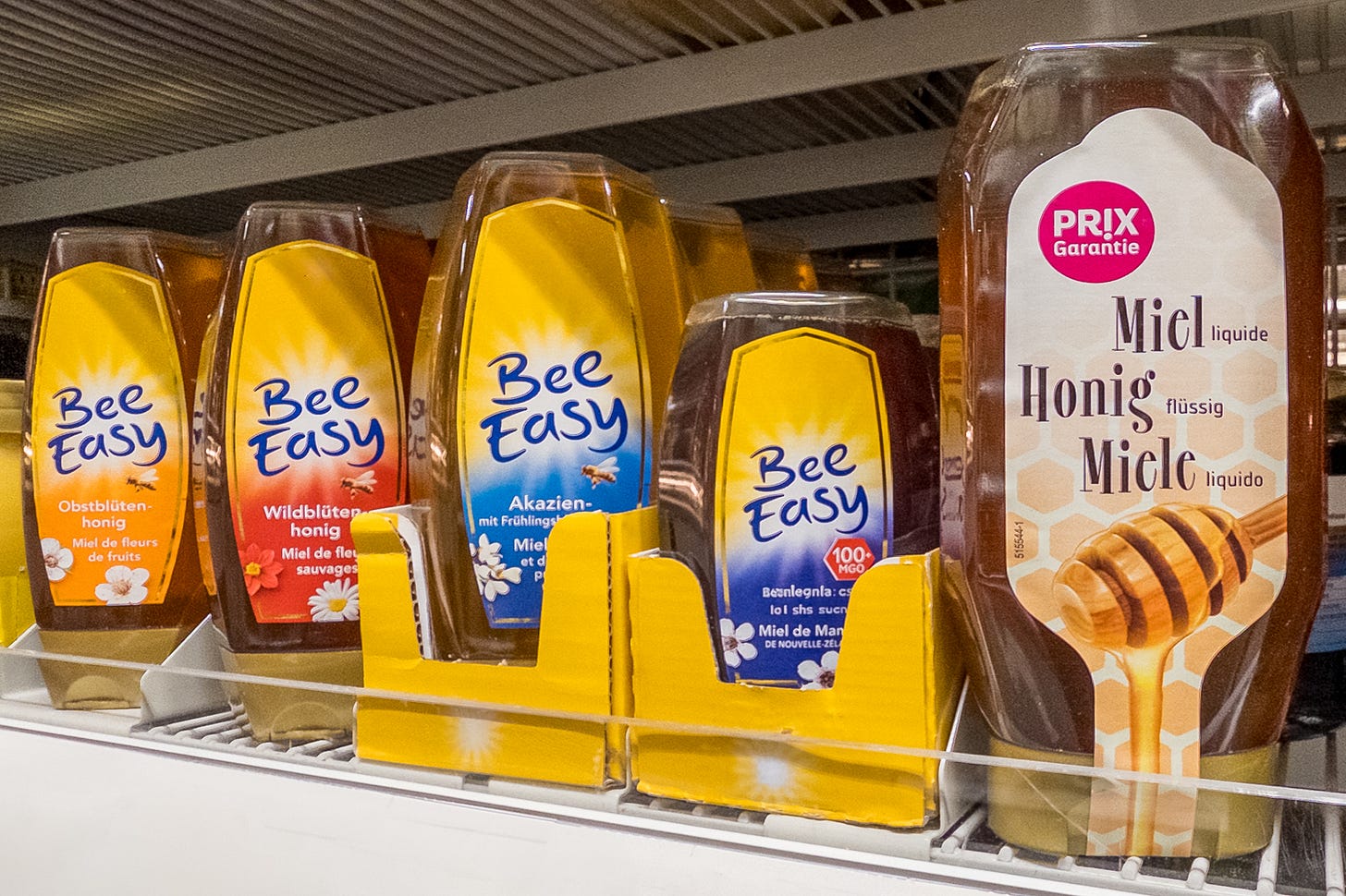 Das Bild zeigt verschiedene Importhonige im Regal eines Schweizer Supermarktes. Die Marken wurden weg retuschiert. Die Sorten umfassen Obstblütenhonig, Wildblütenhonig, Akazienhonig, Manuka-Honig – alles Importhonige, die in transparenten Plastikspendern und Gläsern präsentiert werden. Das Bild zeigt verschiedene Importhonige im Regal eines Schweizer Supermarktes. Die Marken wurden weg retuschiert. Die Sorten umfassen Obstblütenhonig, Wildblütenhonig, Akazienhonig, Manuka-Honig – alles Importhonige, die in transparenten Plastikspendern und Gläsern präsentiert werden.