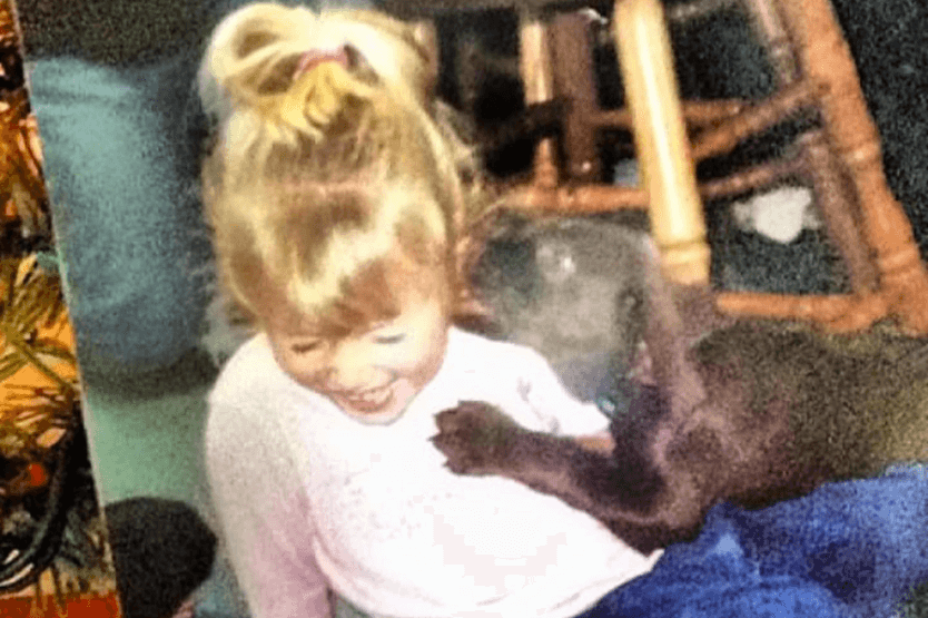 An old disposable camera photo of Haley as a toddler being licked by a chocolate lab puppy