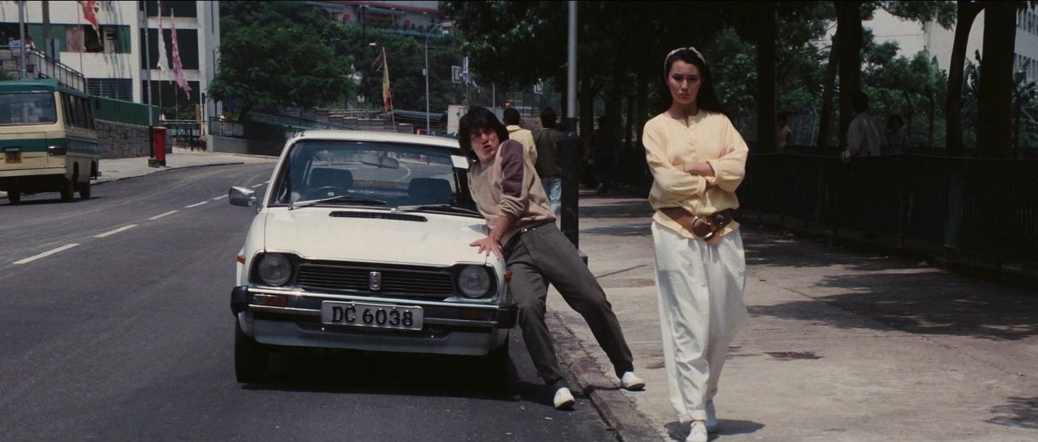 A man is trying to keep a small white car from rolling forward while a woman stands with her arms crossed looking annoyed.