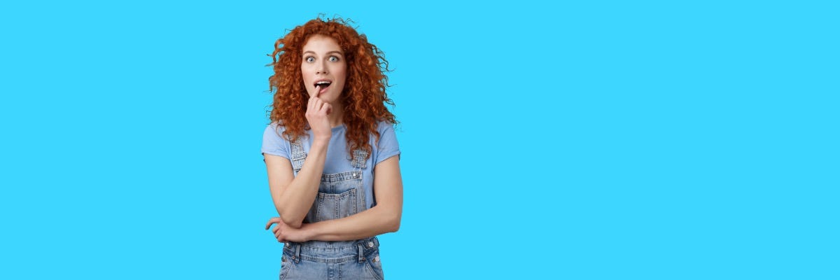 Surprised woman with curly red hair in overalls, touching her chin, against a blue background.