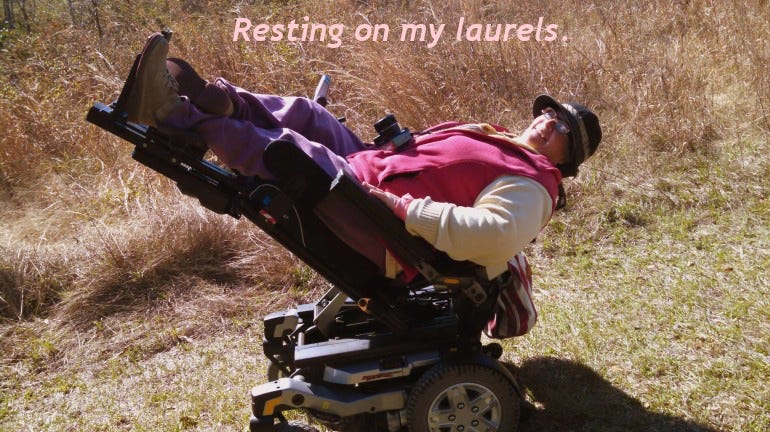 A white woman in a power chair that is tilted way back with the foot rests raised high. She is perched on a grassy path. The text says "Resting on my laurels."