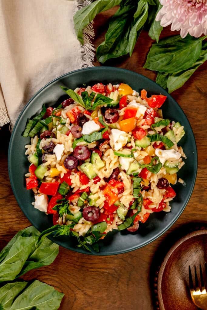 A big green bowl of Greek orzo pasta salad on a wood table surrounded by fresh basil leaves and fresh flowers.