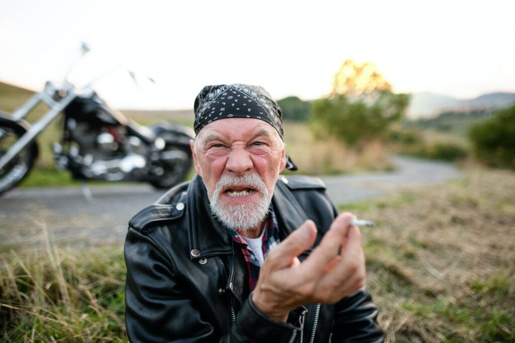Angry senior man traveller with motorbike in countryside, smoking.