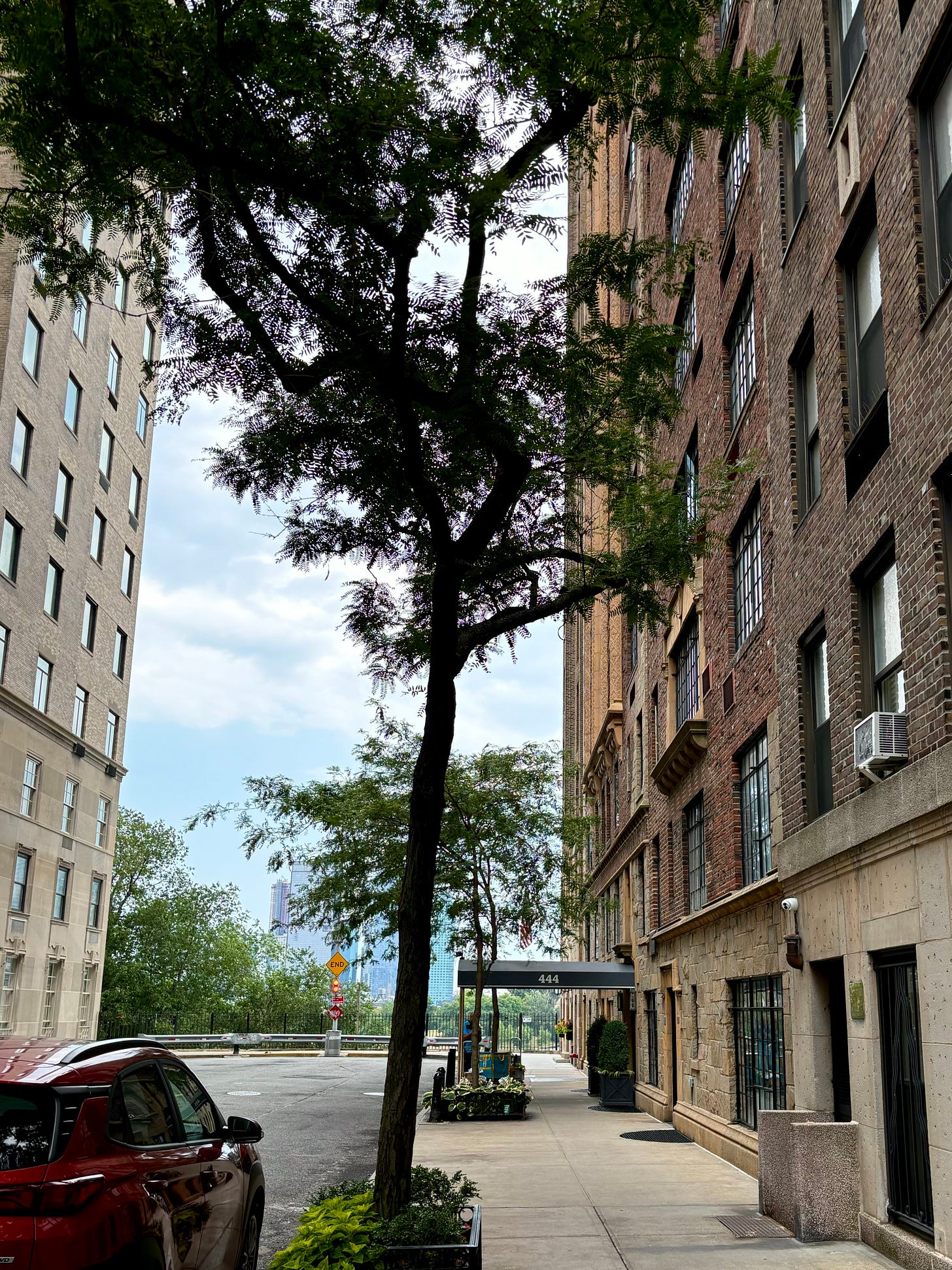 An art deco style red brick apartment building. There is a tree on the sidewalk and the black awning is marked 444. The street is a cul-de-sac, and the East River is beyond the end of the road.