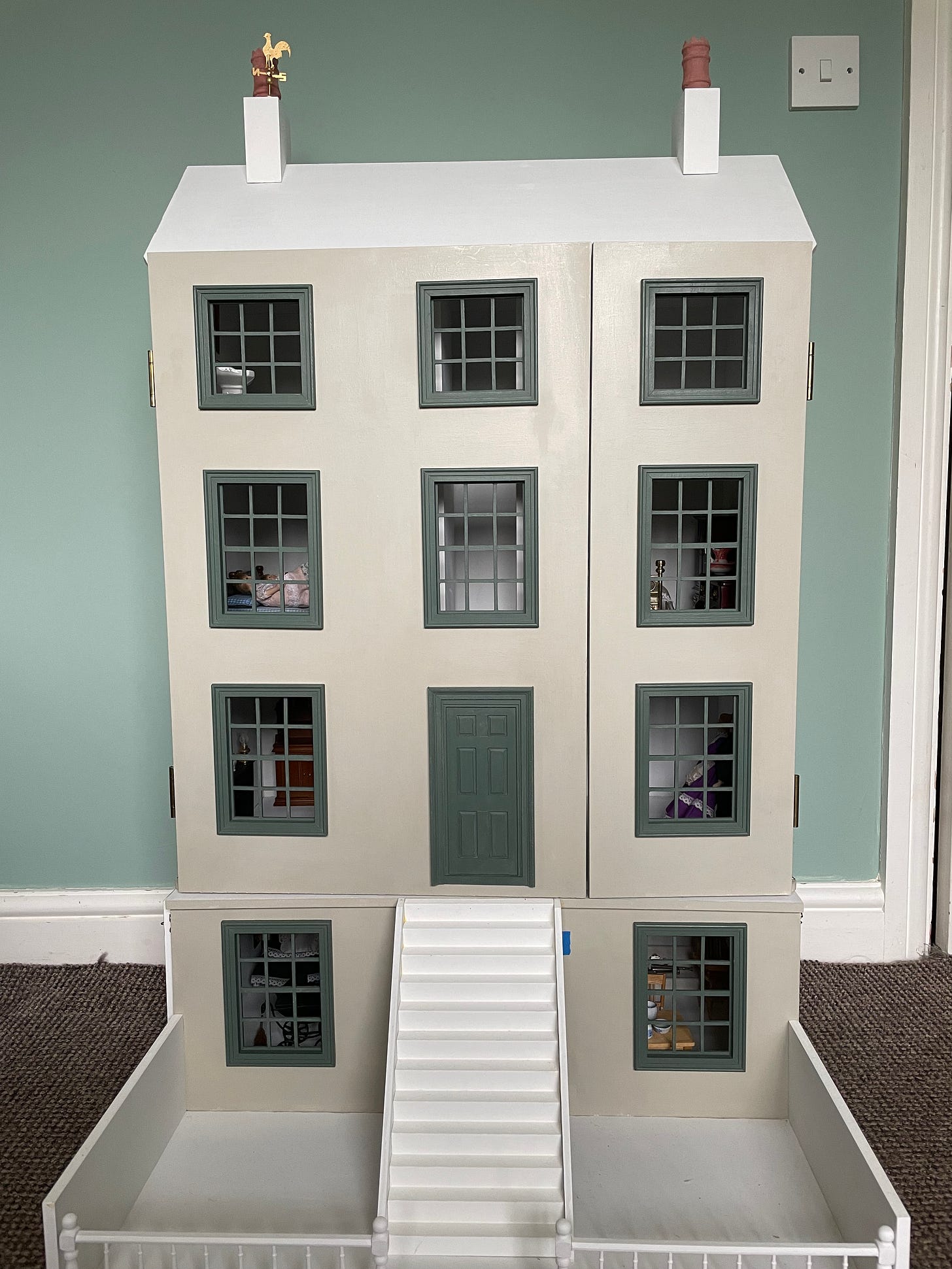 A doll's house in progress. It has a central front door, ten windows, steps up to the front door and a courtyard garden at the front. The walls and windows are painted in two different shades of green, but the roof, steps and garden are all as yet unpainted.