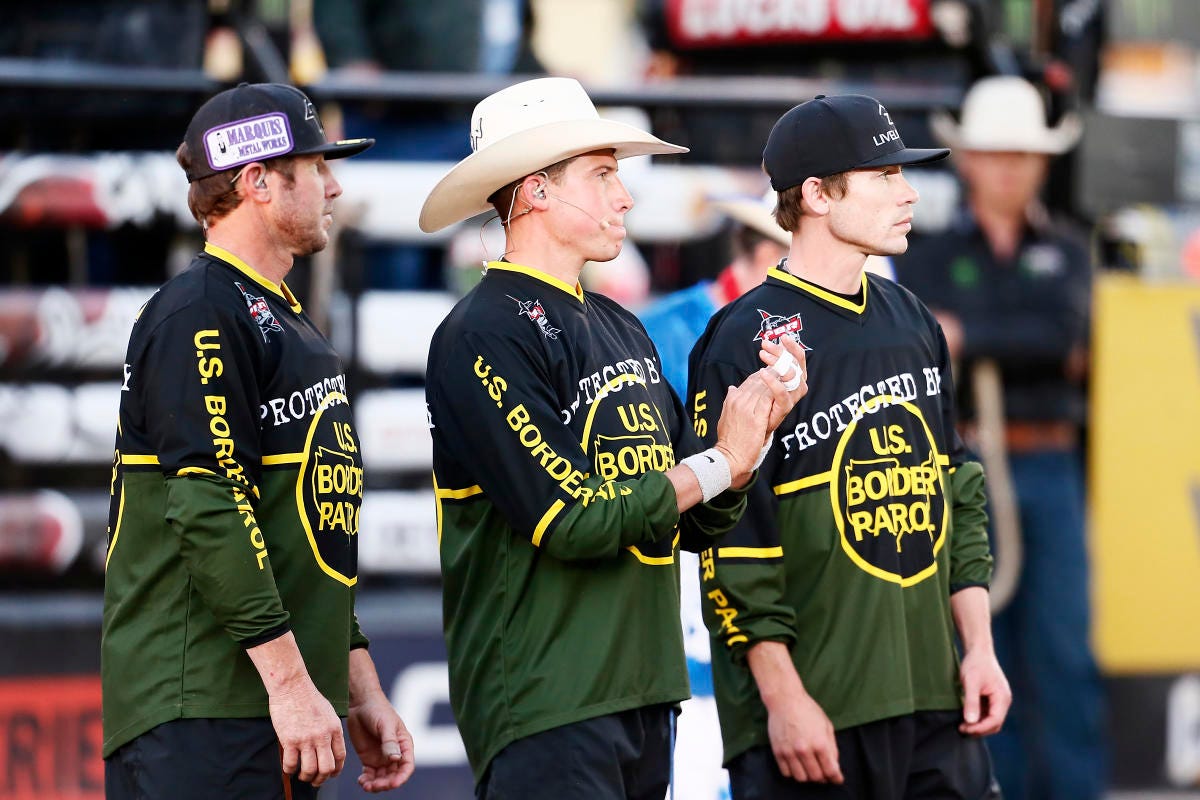 Bull-handlers for the Professional Bull Riders stand in a bullring wearing jerseys sponsored by US Border Patrol.