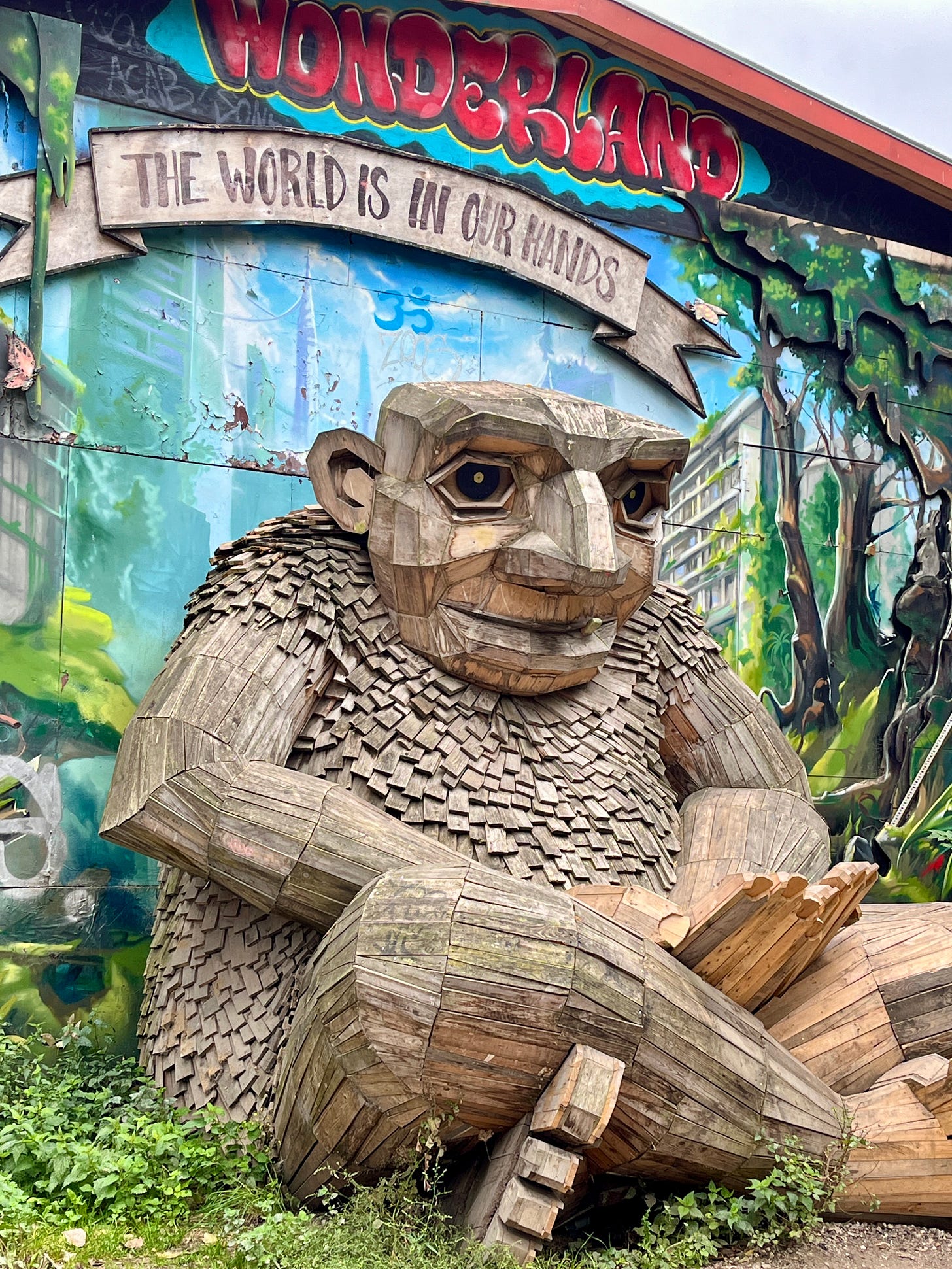 Giant wooden troll made of reclaimed wood, including shingles all over his body. The troll is sitting cross-legged in front of a mural that says "Wonderland" in red, and then "The World is in our hands." The troll's facial expression is neutral enough to be interpreted as the viewer wishes.