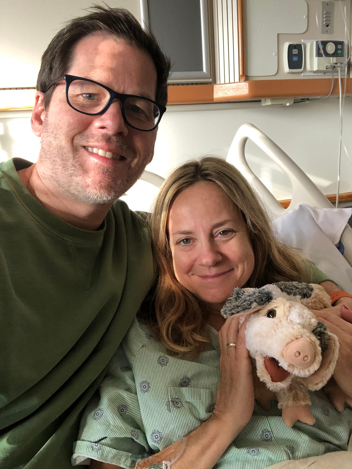 Memoir coach Christine Wolf with her boyfriend, Eric Ronne, after her surgery. They're sitting together in a hospital bed, holding a stuffed animal, smiling. 
