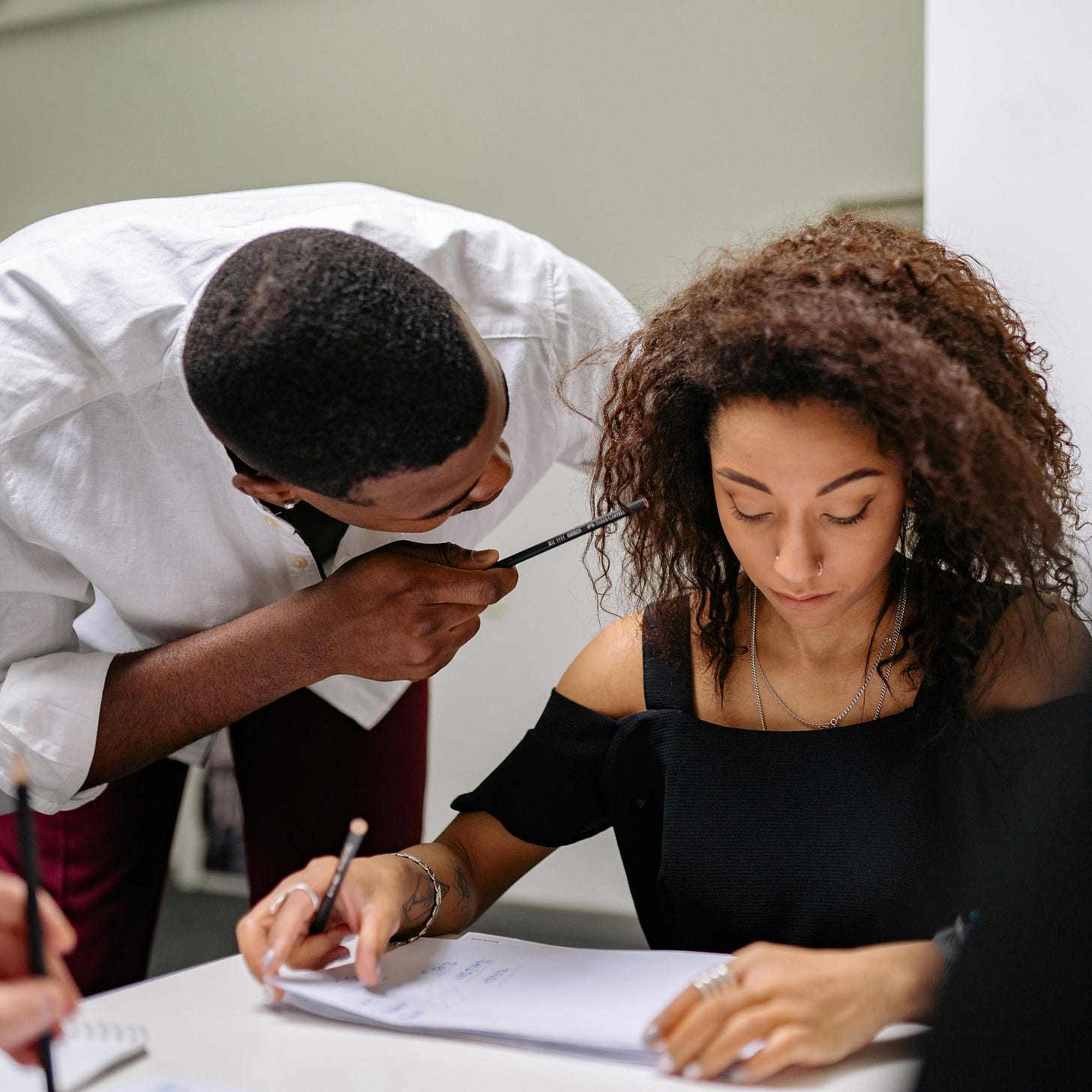 Photo of man in white shirt getting close to a woman seated at a desk and pointing a pencil in her face 