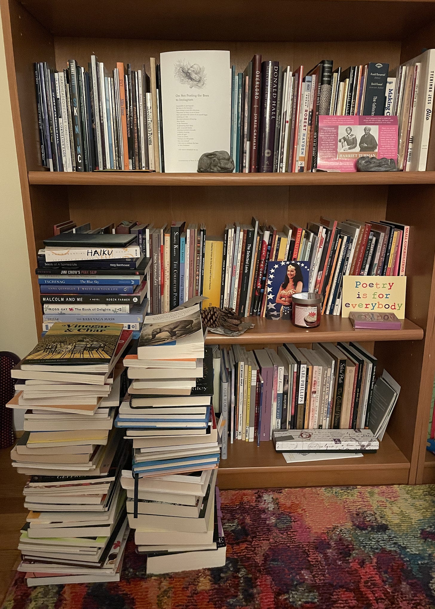 three shelves of a bookcase with two stacks of books on floor in front that reach to 2nd shelf. Also visible on bookcase is an image of Wonder Woman, a postcard that reads "Poetry is for Everybody," a postcard of Harriet Tubman quotes, a broadside of a poem, and various knick knacks.