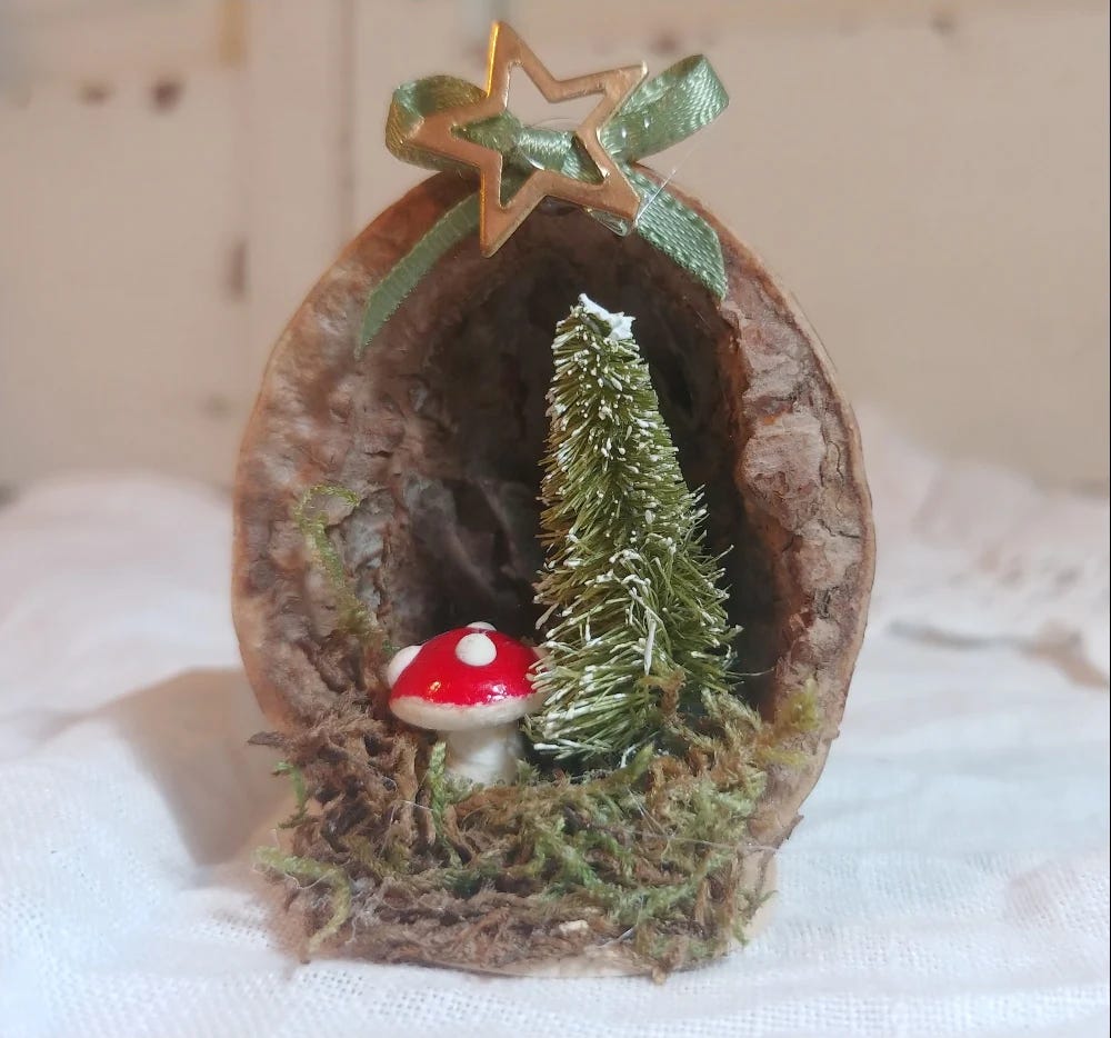 A small diorama-style Christmas ornament made of a tiny tree and mushroom inside of a walnut shell, with a green ribbon attached.