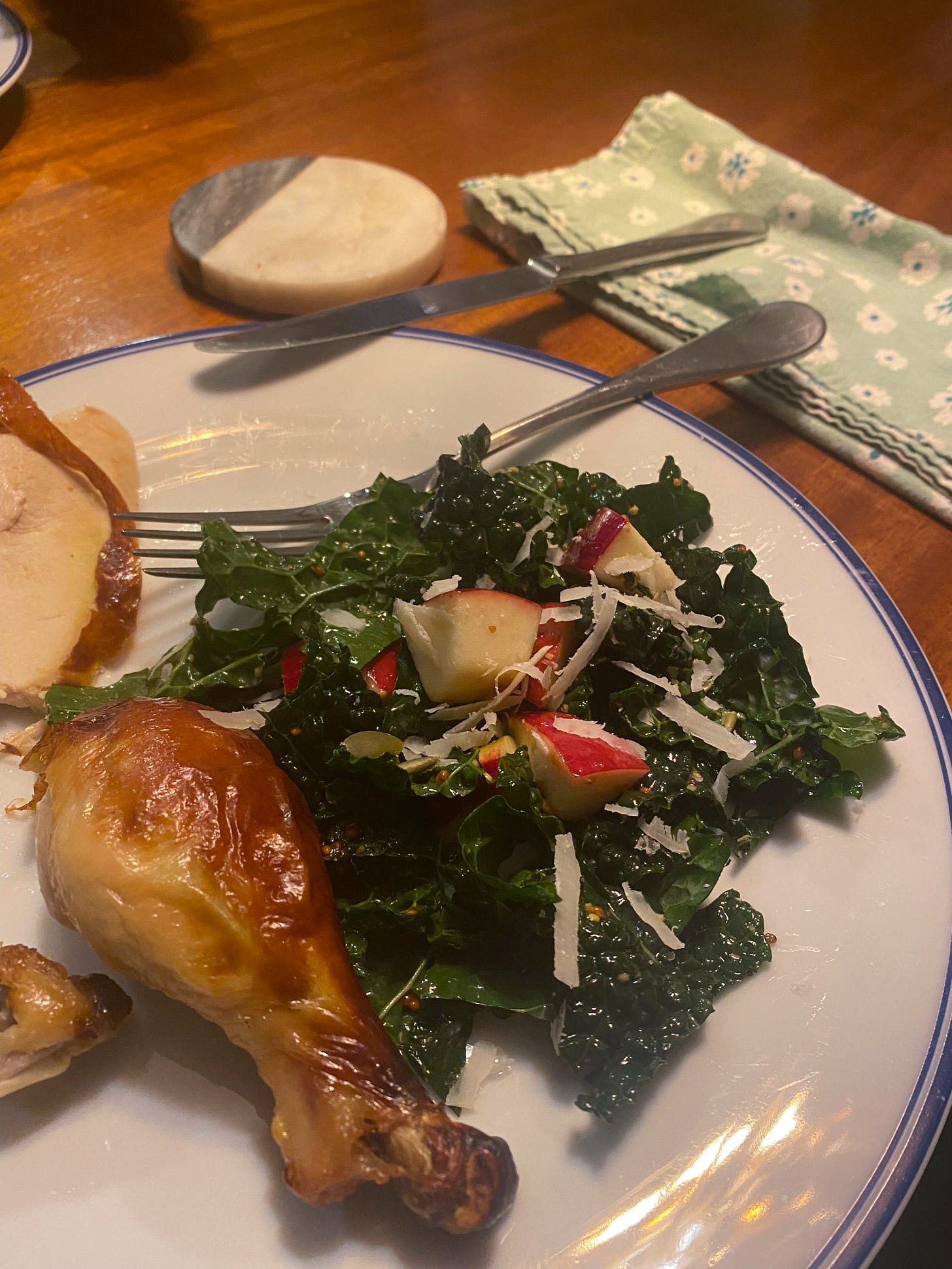 On a white dinner plate with a blue rim is a chicken drumstick and a bit more of the meat visible at the left side of the frame. To the right of the chicken is a lacinato kale salad with red-skinned apple pieces, pepitas, and coarsely grated pecorino.