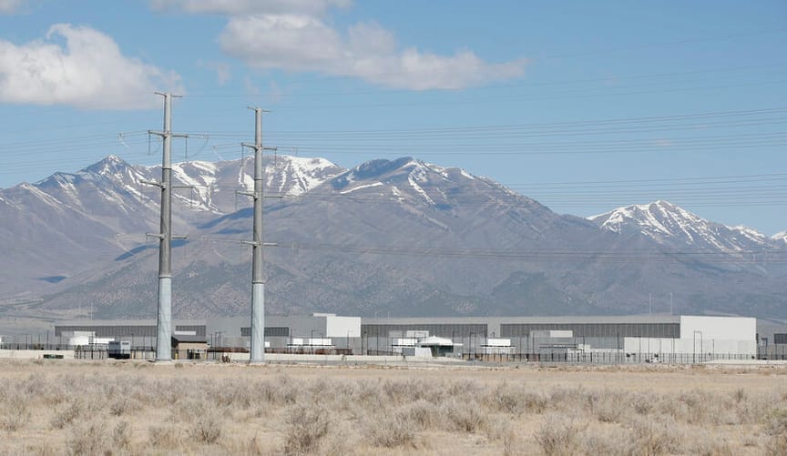 A large industrial facility with two transmission towers seen in the foreground. Snow-capped mountains stand at rear.