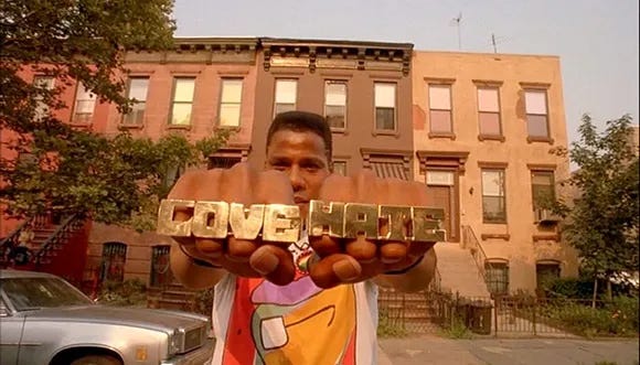 This image shows the Do the Right Thing character of Radio Raheem (played by Bill Nunn) looking directly into the camera as he holds both hands in front of him. On his right hand, Raheem wears a four-finger golden ring in the shape of the word "LOVE." On his left hand, Raheem wears a four-finger golden ring in the shape of the word "HATE."