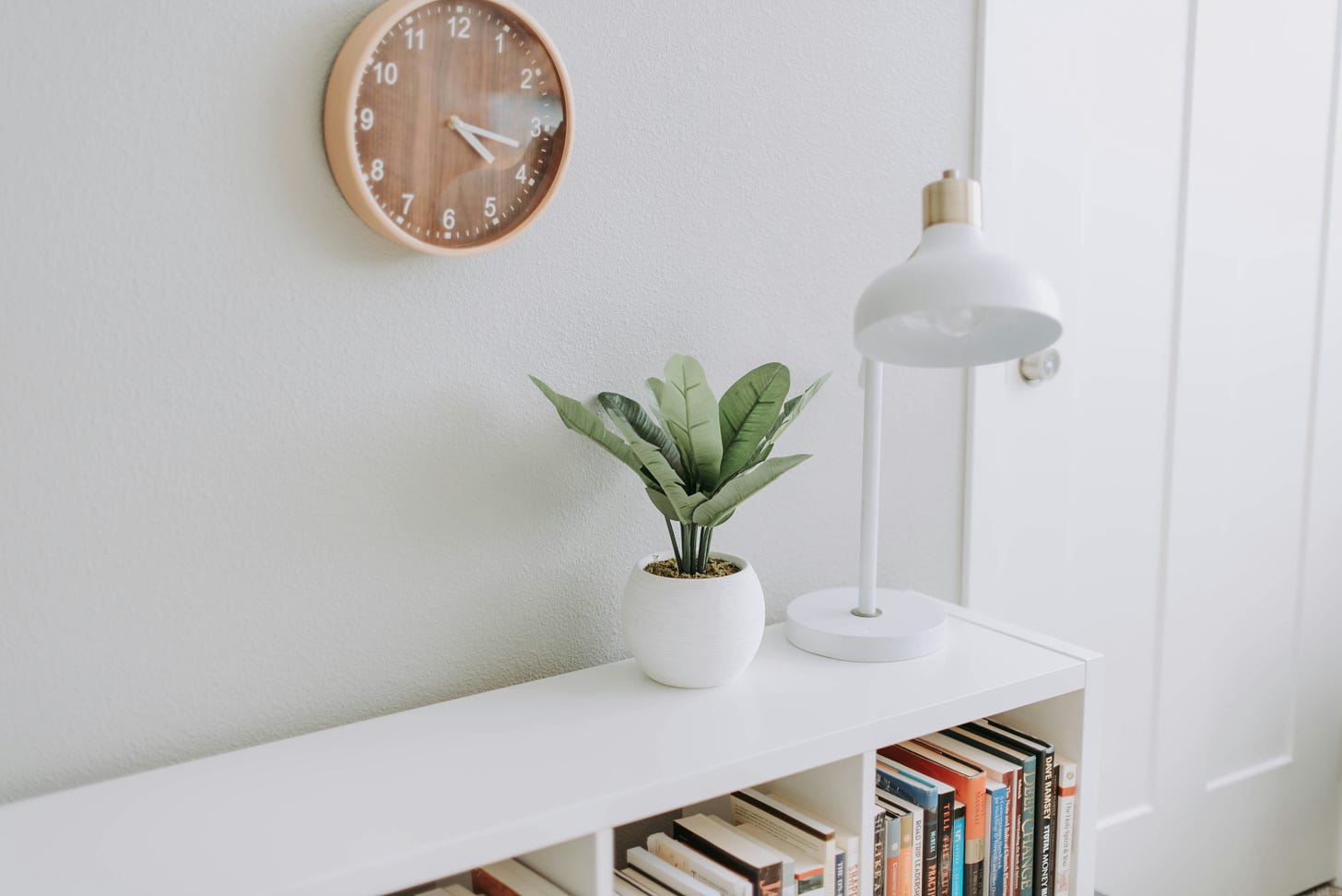 a clean, minimal, organized room. all walls and surfaces are painted white. a small green plant in a white planting pot, and a white lamp sit on top of a bookshelf. the bookshelf is filled with colorful books. a brown analog clock hangs above on the wall