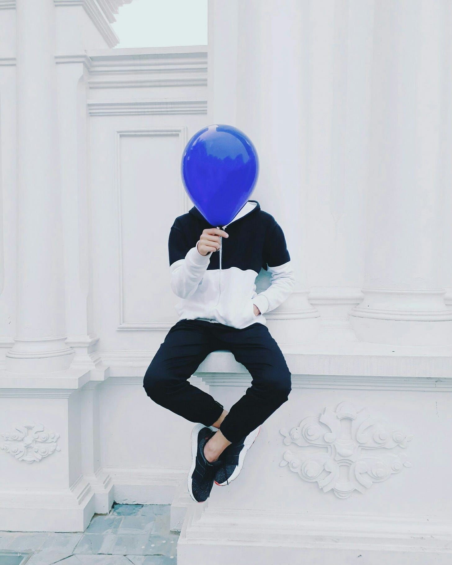 A person in black pants and a white and black block-print sweatshirt, holding a royal blue balloon in front of their head. 
