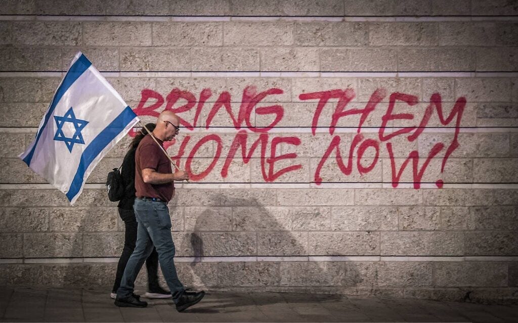 An Israeli couple holding an Israeli flag in Jerusalem, walk in front of graffiti calling for the release of Israeli hostages held in the Gaza Strip since October 7 2023 by Hamas (Gil Cohen-Magen / AFP)