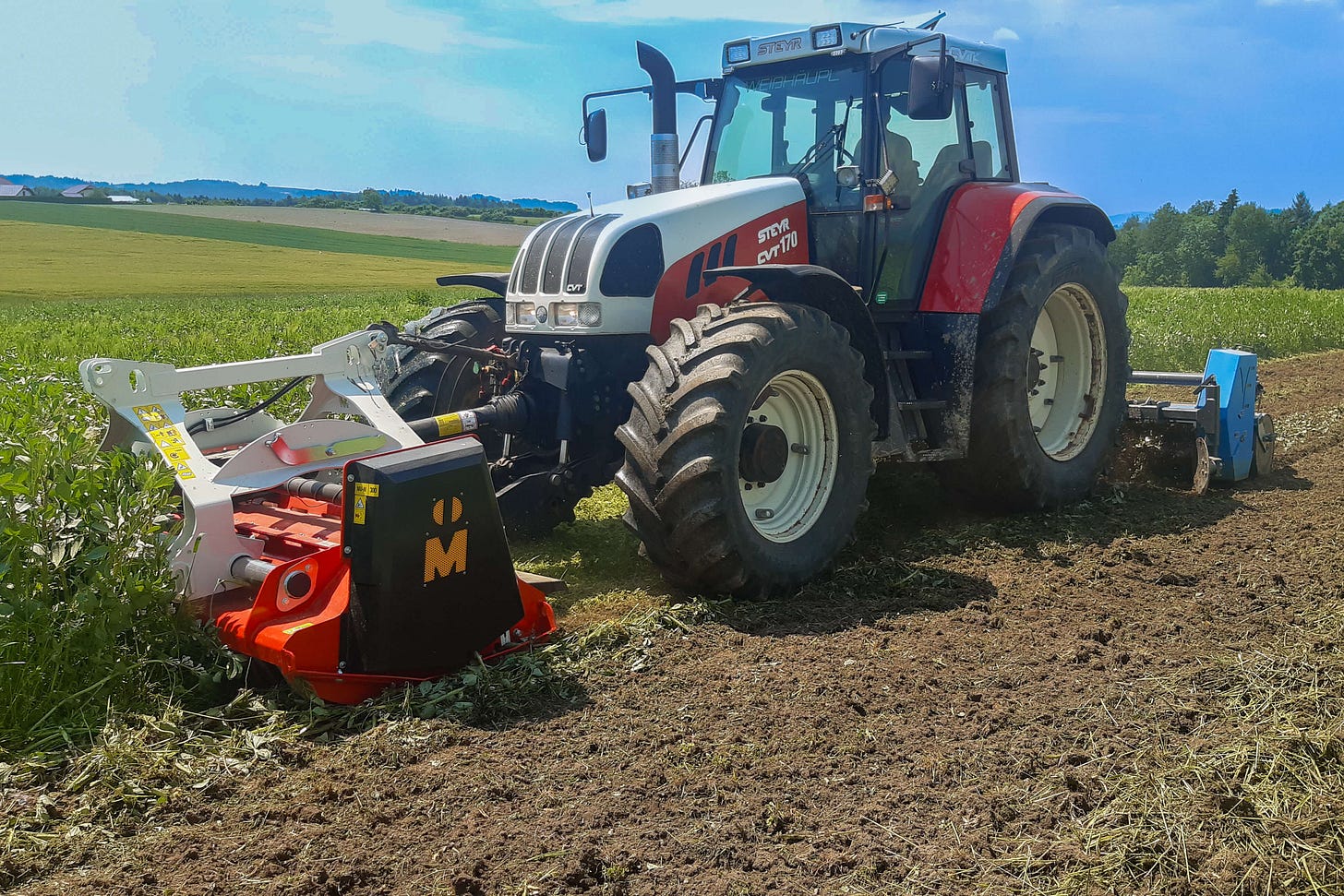 Ein Steyr-Traktor mit Frontmähwerk mäht und häckselt eine wüchsige Zwischenfrucht auf einem Acker. Die zerkleinerte Pflanzenmasse wird hinter dem Traktor mit einer Fräse eingearbeitet – ein wichtiger Schritt in der regenerativen Landwirtschaft zur Förderung des Bodenlebens. Ein Steyr-Traktor mit Frontmähwerk mäht und häckselt eine wüchsige Zwischenfrucht auf einem Acker. Die zerkleinerte Pflanzenmasse wird hinter dem Traktor mit einer Fräse eingearbeitet – ein wichtiger Schritt in der regenerativen Landwirtschaft zur Förderung des Bodenlebens.