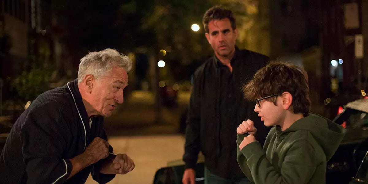 Promotional photo from the movie Ezra. Robert DeNiro and William A. Fitzgerald, a young white actor with shaggy brown hair and glasses, are playing at holding their fists up at each other. Actor Bobby Cannavale, a white man with curly black hair, looks on from the background with unease.]