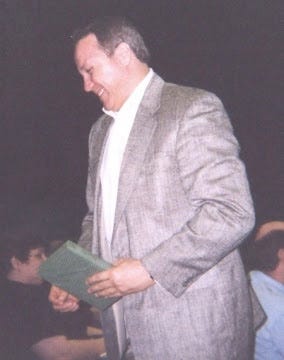 Peter Christopher, holding a book and smiling at someone in an auditorium as he walks up an aisle wearing a gray suit and holding a green book