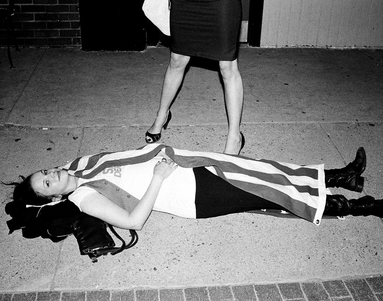 Amber lying on a sidewalk. She looks to camera, smiling. Behind her, there is a woman's legs. The woman is wearing a skirt and high heels. The upper half of her body is out of frame. 