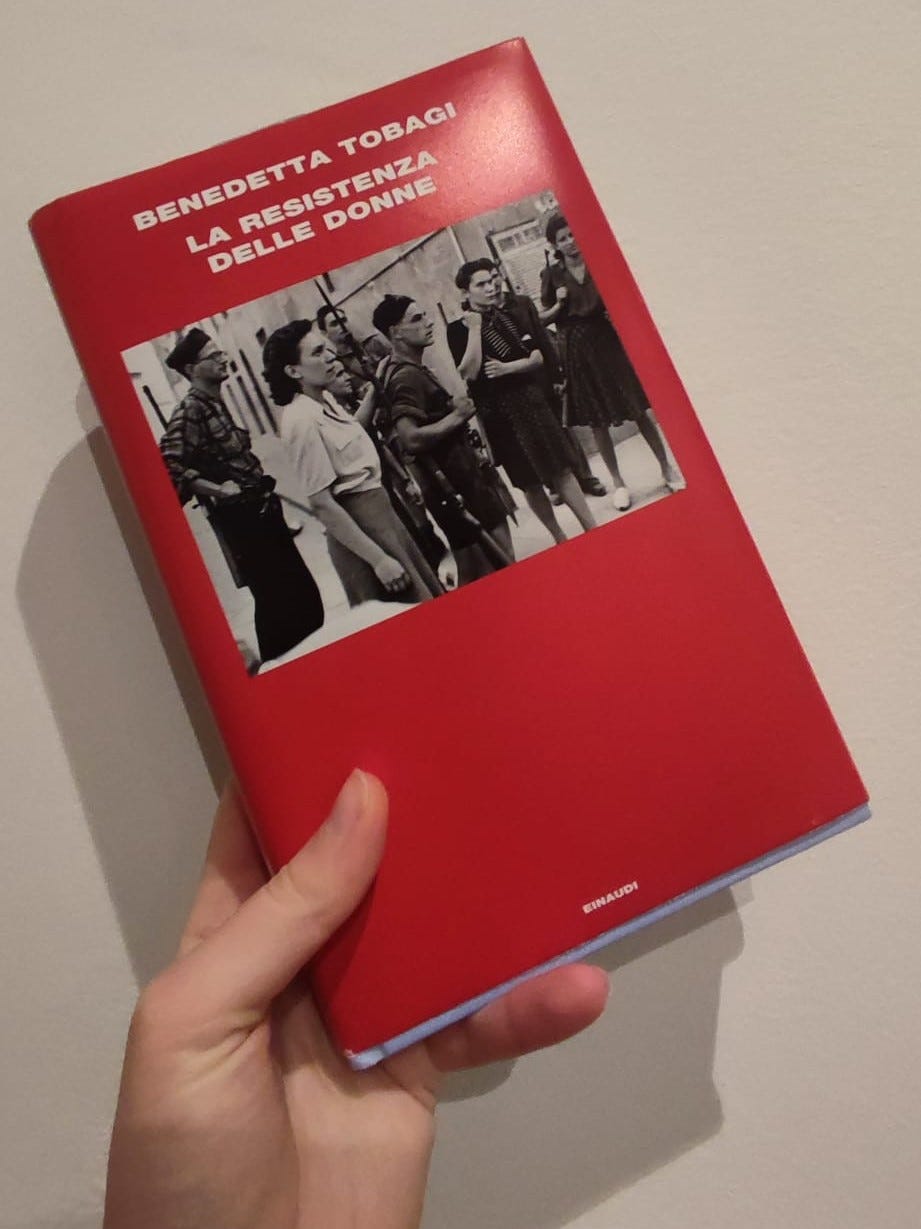 Una mano tiene un libro rosso con una fotografia di partigiani nella parte alta. Dietro al libro, un muro bianco