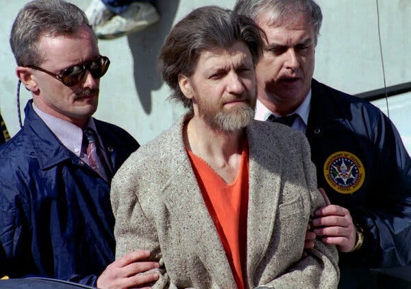 Theodore Kaczynski, shaggy-haired and bearded and wearing an ill-fitting gray sports jacket over an orange prison uniform, with federal agents on each side holding onto his arms.