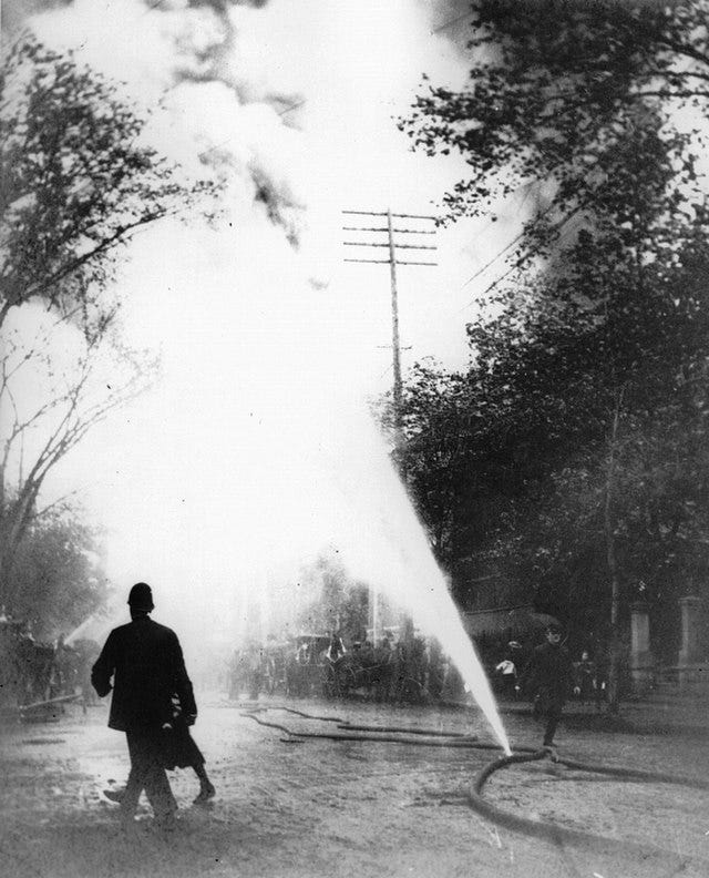 black and white photograph of city street with fire hose bursting water out