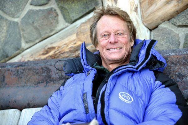 Man in blue snowsuit sitting against wall and smiling to camera.