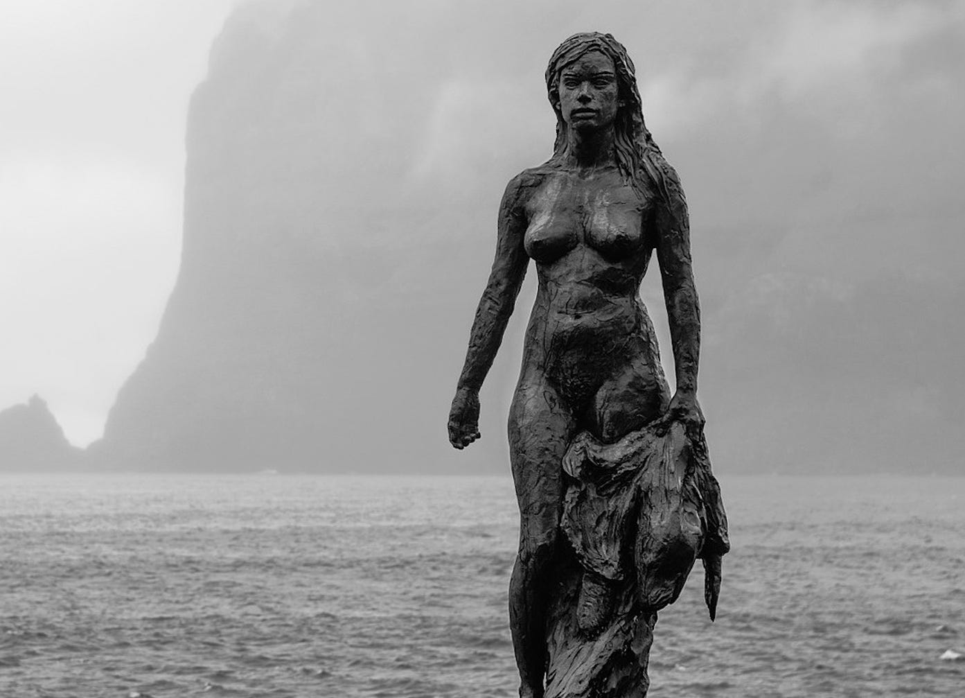 A bronze statue of a naked woman holding a seal pelt stands in a grey sea mist, with the sea behind her and imposing cliffs in the background A bronze statue of a naked woman holding a seal pelt stands in a grey sea mist, with the sea behind her and imposing cliffs in the background