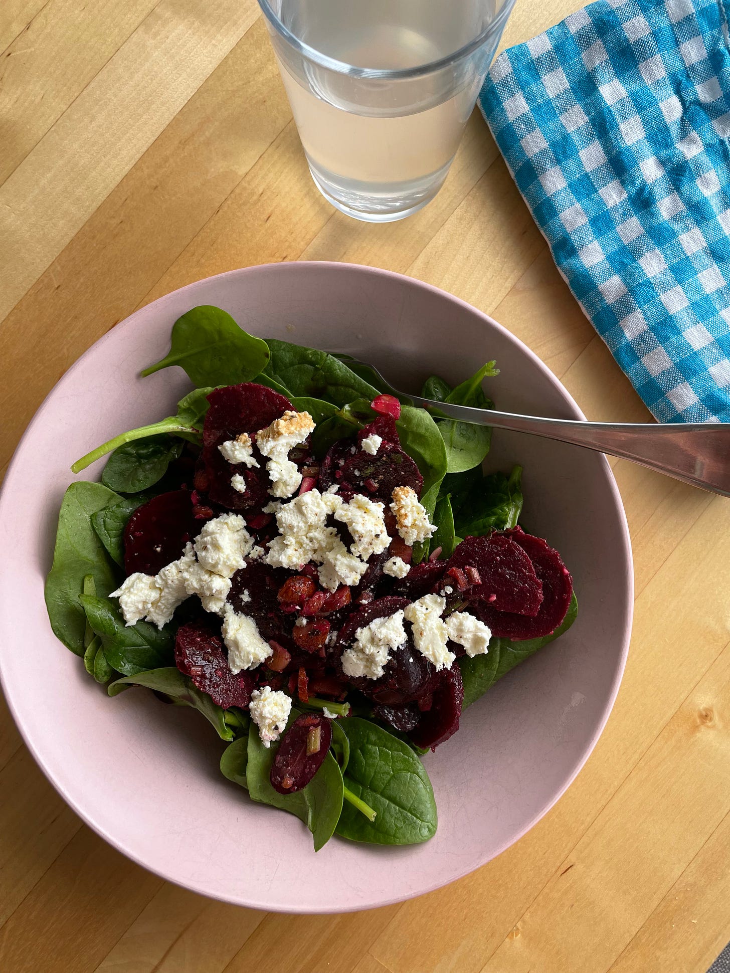 Pink bowl filled with baby spinach leaves, chopped cooked beetroot, feta cheese, parsley vinegarette and roasted almonds. A glass of coconut water and chequered napkin nearby.
