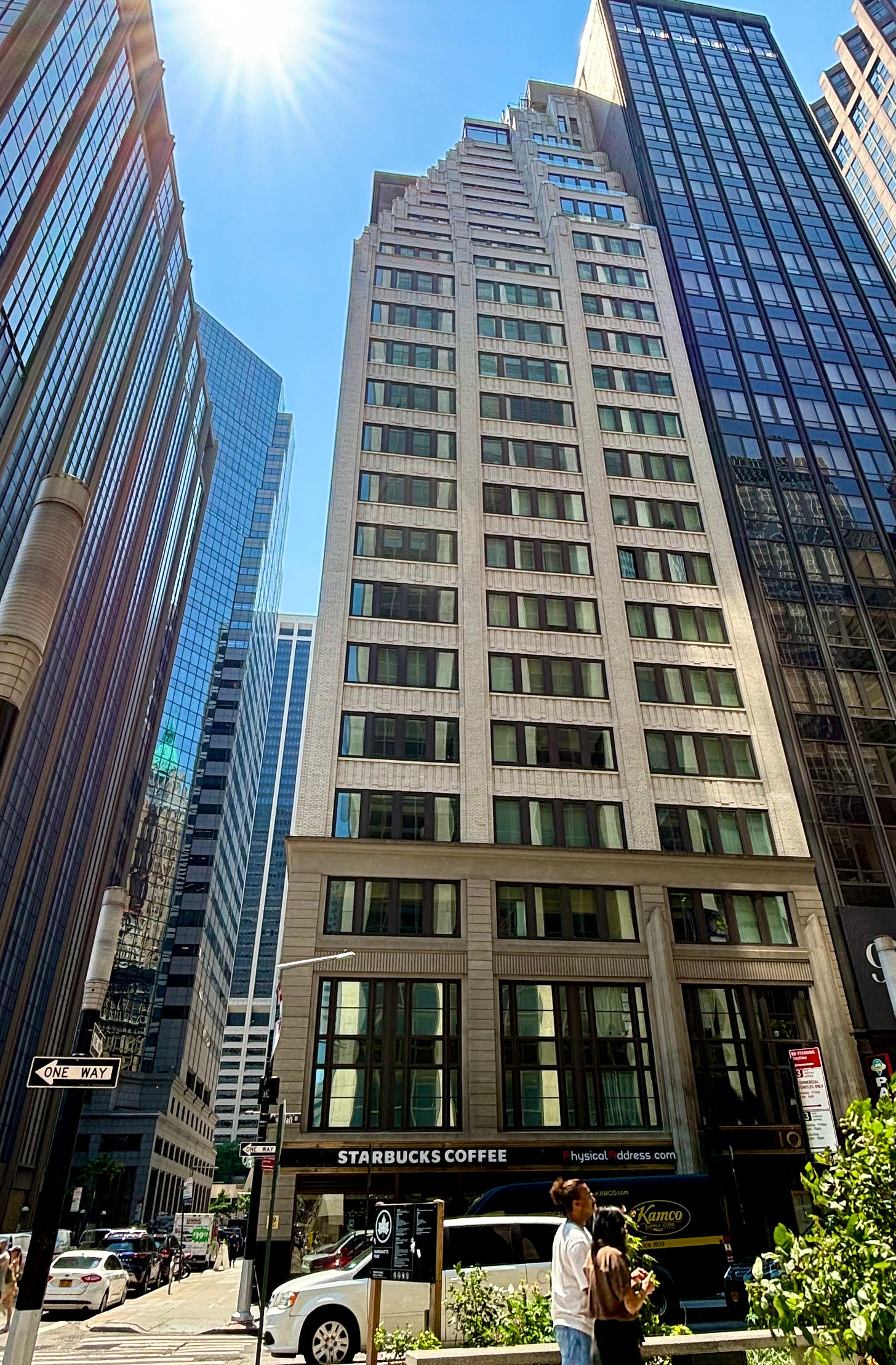 A tall building with a small 99 on the storefront level. The upper floors are stepped back. The sky is blue and the sun is intensely shining. There is a Starbucks.