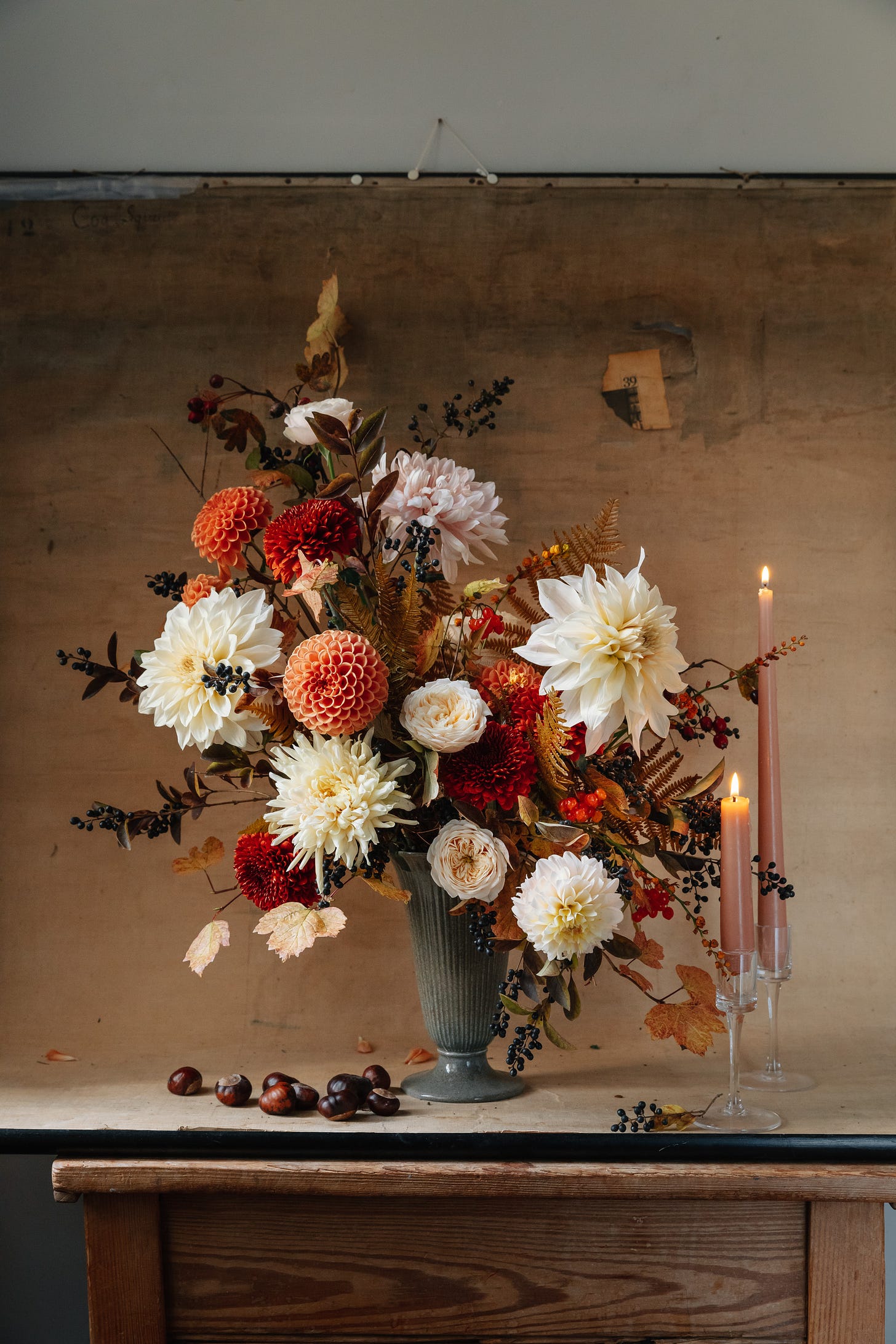 An arrangement of autumn flowers in a tall vase, two lit candles to the right of the flowers and placed on top of a small table. The background is a roll of worn tattered canvas.