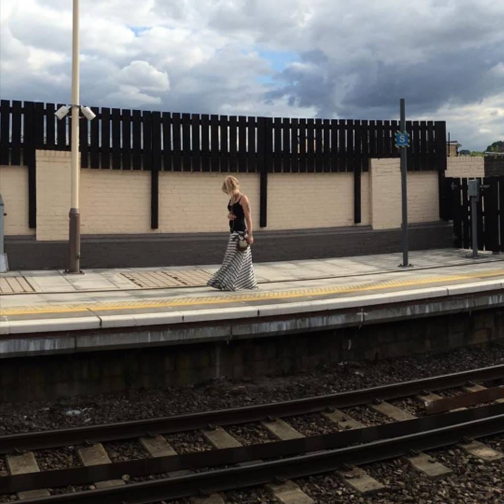 Overground train station platform in London, summer time, showing a young blonde woman on the other side of the tracks, looking down at her feet. The sky is overcast.