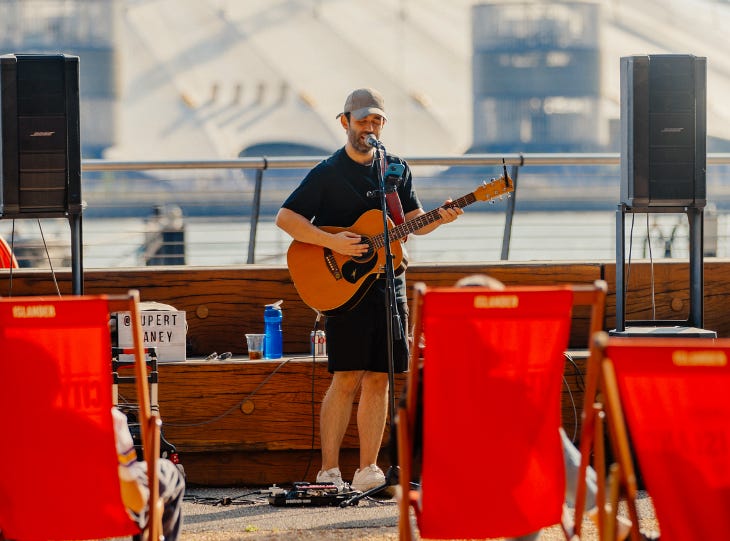 A musician singing and playing guitar to an audience sitting in deckchairs, in front of the O2