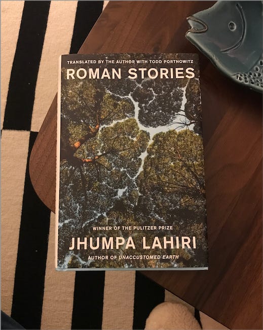 Lahiri's book, which has trees on the cover, rests on a wooden table next to a dish shaped like a fish. Below the table is a black-and-white striped rug and a pair of fluffy slippers.