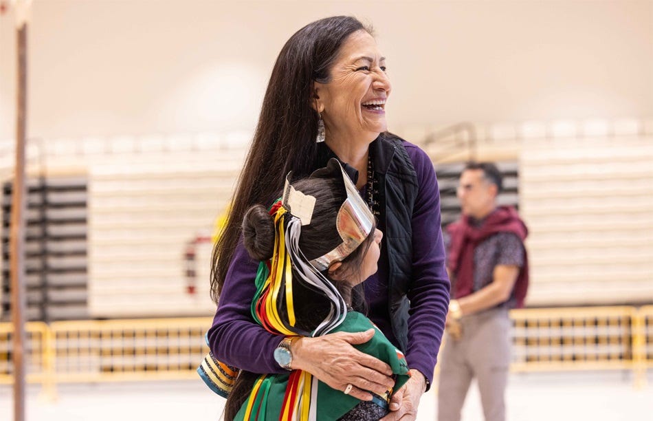 In an undated photo, then-Interior Secretary Deb Haaland smiles as she hugs a girl wearing a green cape and headdress with bright yellow, red, green, and white ribbons In an undated photo, then-Interior Secretary Deb Haaland smiles as she hugs a girl wearing a green cape and headdress with bright yellow, red, green, and white ribbons