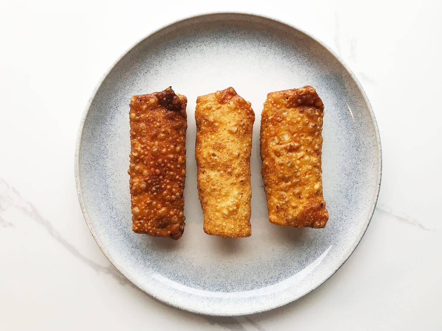 Three bubbly, deep fried egg rolls on a plate. The one on the left is well done (but not burnt!). The one in the middle is a light golden. On the right is medium golden.