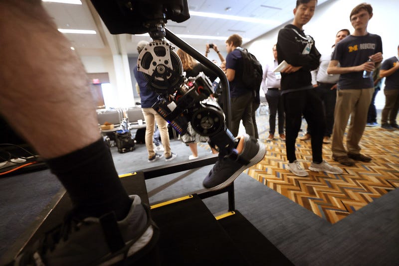 A powered prosthetic knee-ankle with a large gear for the knee is raised midway up a small staircase in a conference setting as a small crowd observes in the background.