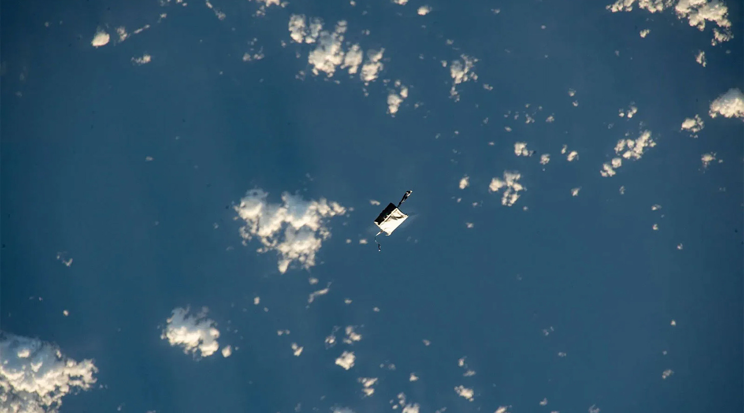 A photo (from above) of a white fabric tool bag floating above the surface of the Earth, which appears as a blue background with small fluffy white clouds 
