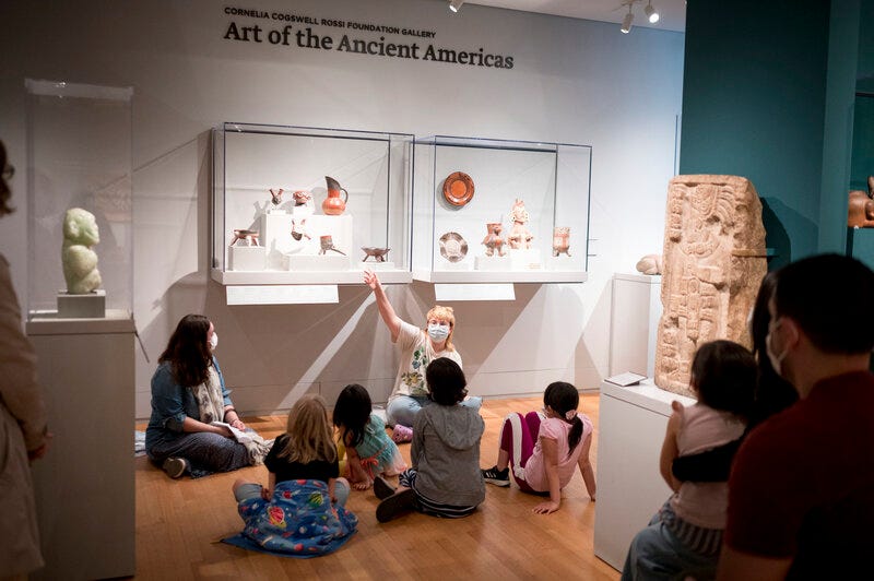 A group of children seen from behind as they sit on the floor of a gallery. They look toward a seated adult, who wears a surgical mask and gestures toward a vitrine full of objects, including a pitcher. 