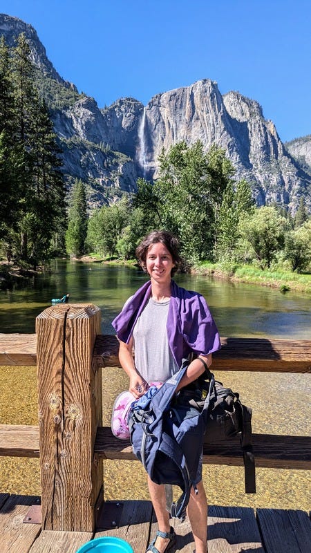 Rey, a white nonbinary person with short brown hair, stands smiling on the bridge with a large waterfall in the distance. The river we floated down is also in the background.