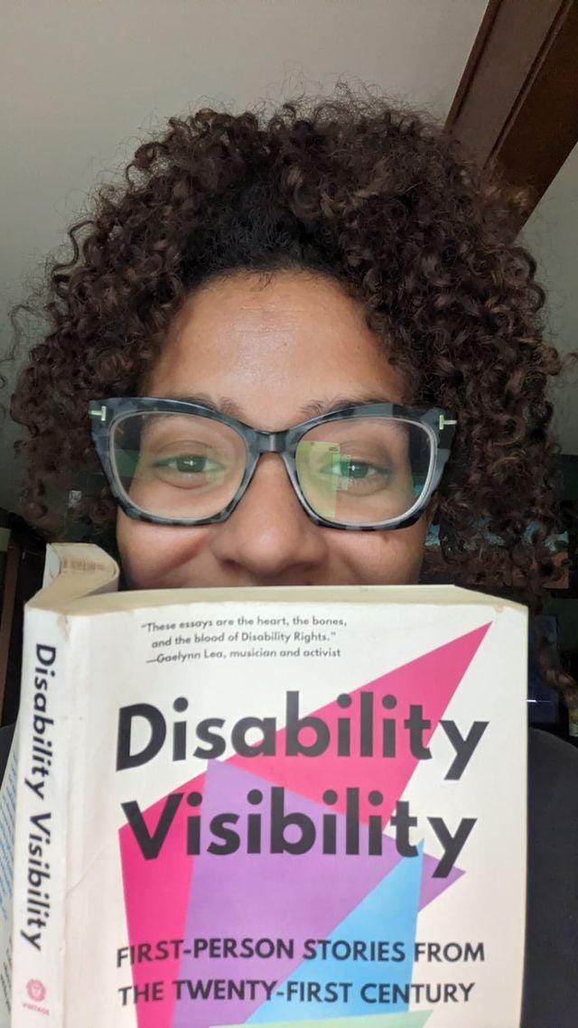 A person wearing glasses holding a book titled "Disability Visibility" up to their face so that only the top half of their face is visible