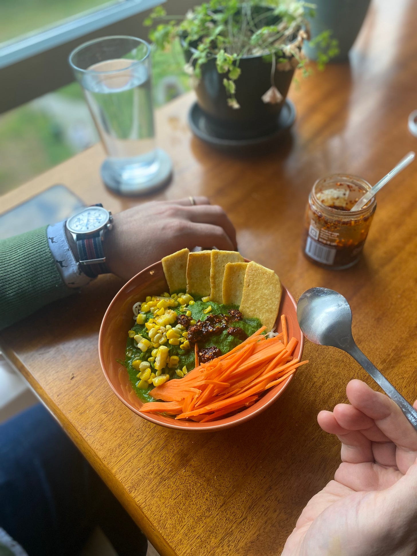 Jeff's hands are at either side of an orange bowl full of ramen noodles in a green sauce, topped with carrot matchsticks, corn, chili crisp, and slices of pan-fried tofu. He's holding a spoon in his right hand in the foreground.